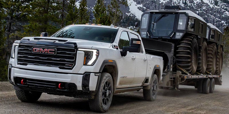 White GMC Sierra 2500HD towing a large off-road vehicle trailer through a rugged mountain landscape